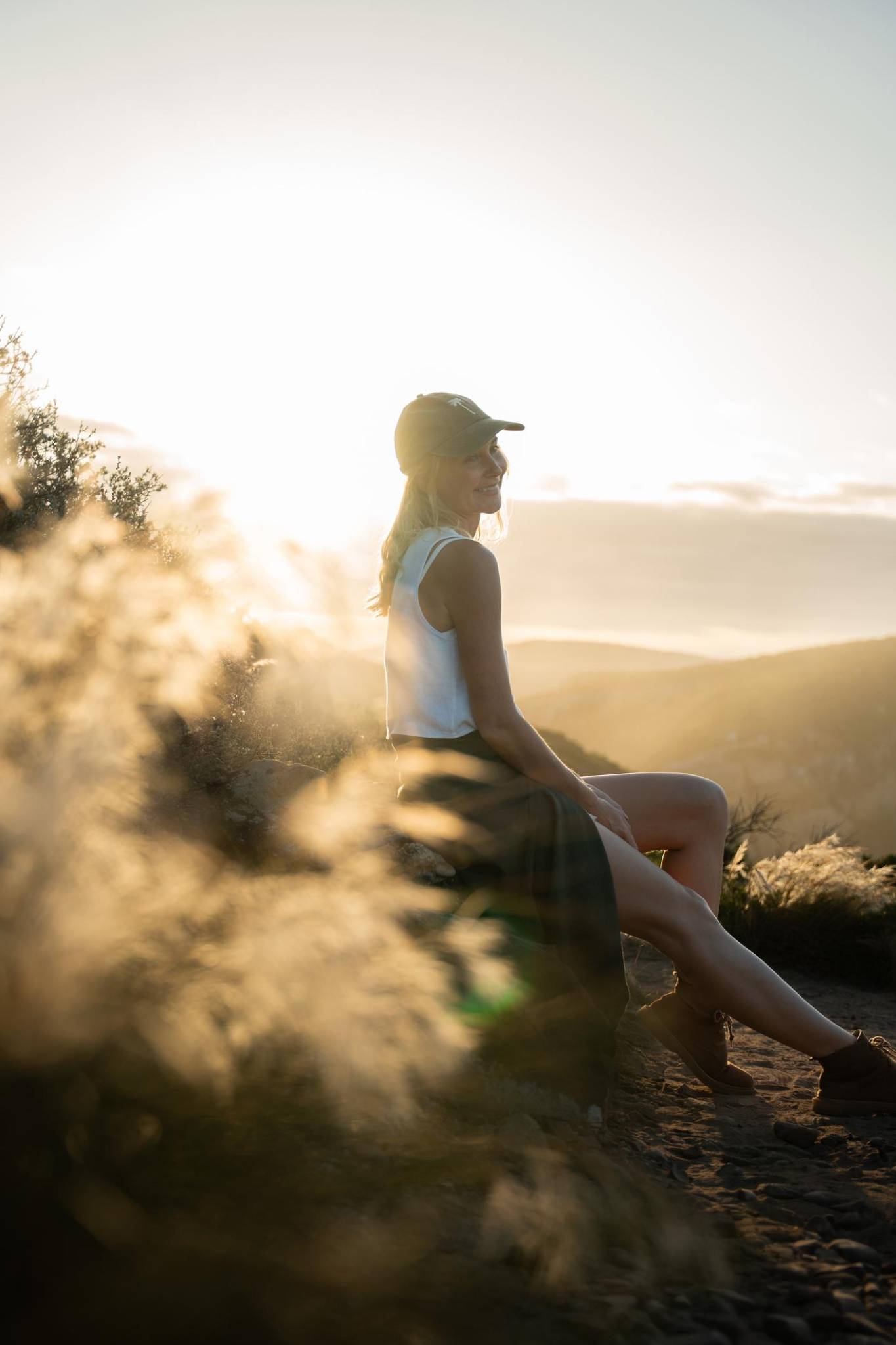 Woman sitting on a mountain trail at sunset, looking out over the landscape in a peaceful, reflective moment