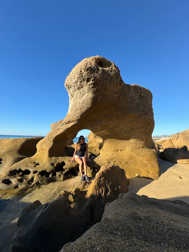 Danielle Alvarez Greer sitting on rocks by the ocean in Cabo San Lucas beneath an elephant-shaped rock formation during a peaceful beach walk