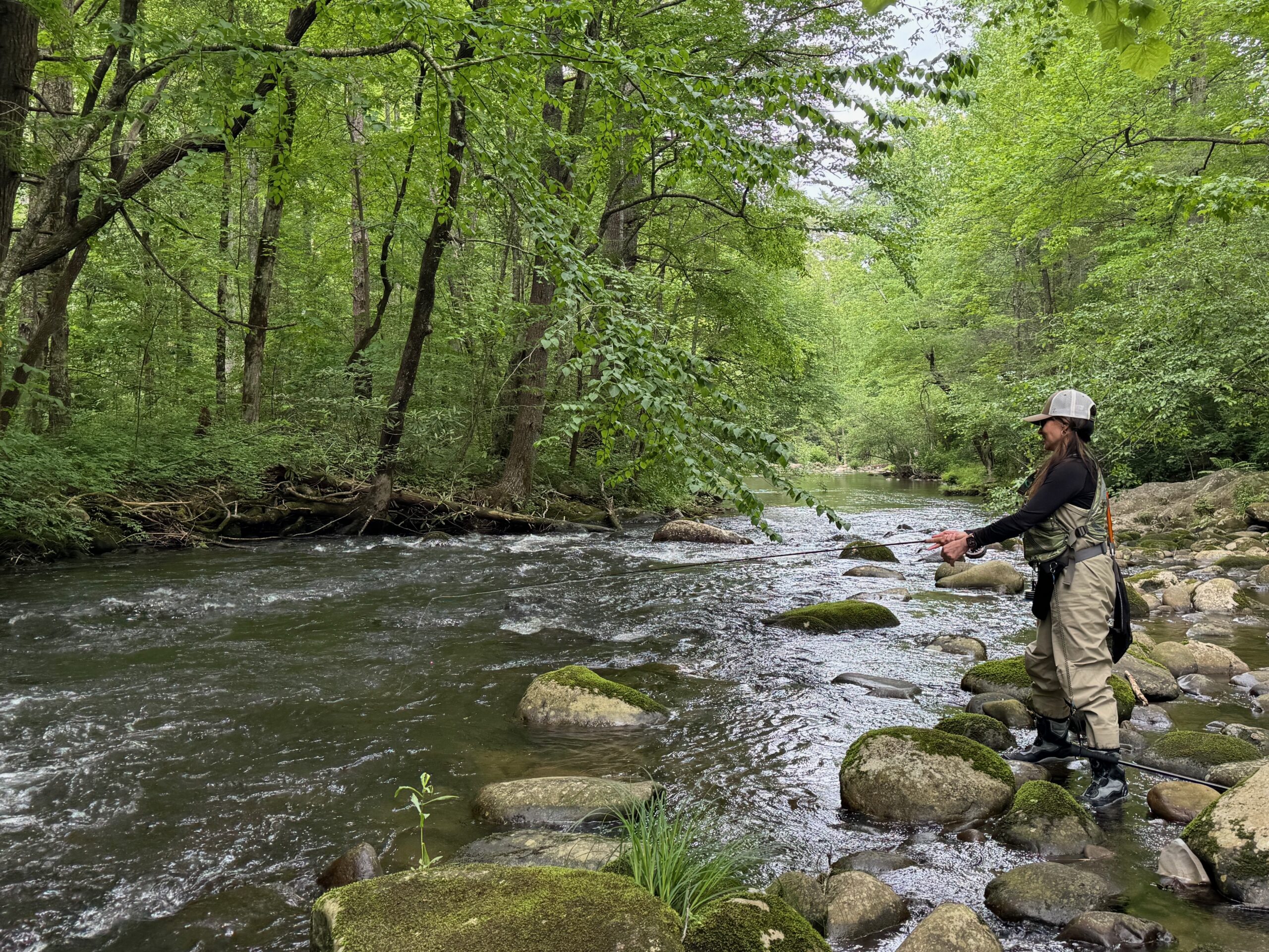 A woman standing in a peaceful river surrounded by trees, quietly reflecting while fishing, symbolizing surrender and trust in God.